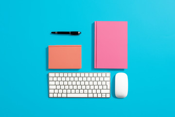 Office equipment on a pastel blue background.  Includes orange and pink hardcover notebooks, computer keyboard and mouse, black pen and black iphone.  Center composition.