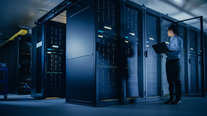 In Data Center: Male IT Technician Running Maintenance Programme on a Laptop, Controls Operational Server Rack Optimal Functioning. Modern High-Tech Telecommunications Operational Super Computer.