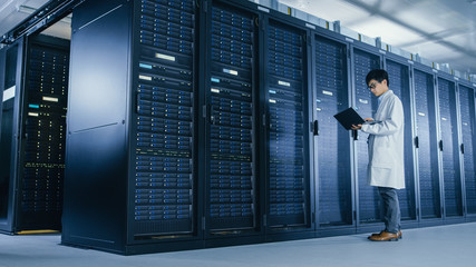 Leinwandbilder - In Data Center: Male IT Specialist Wearing White Coat Stands Beside Server Rack, Uses Laptop Computer to Run Maintenance Diagnostics Tools, Controls so that Database Works at Optimal Level. 