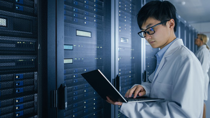 Wall Mural - In Data Center: Male and Female IT Specialists Wearing White Coats Work with Server Racks, Use Laptops to Run Maintenance Diagnostics. People wearing Lab Coats Working with Datacenter Database.