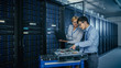© Gorodenkoff - In the Modern Data Center: Engineer and IT Specialist Work with Server Racks, on a Pushcart Equipment for Installing New Hardware. Specialists Doing Maintenance and Diagnostics of the Database.