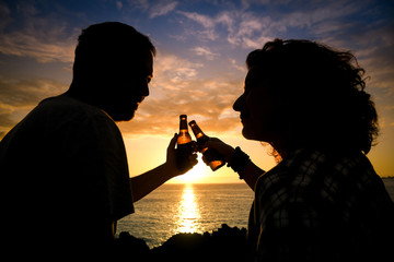 Wall Mural - Happy couple enjoy in beer by the beach during the sunset. Lovers clinking their ale bottles outdoors. Romantic holidays.  Loving two drinking together while resting on the beach enjoying sundown.