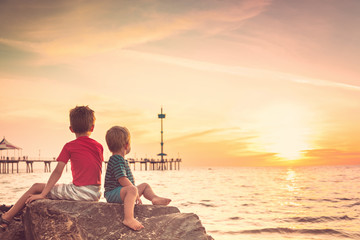 Wall Mural - Two boys sitting on the rock at the beach at sunset