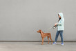 Girl and dog on a leash are walking against the background of a gray wall. Owner walks the dog on a gray background. Street photo of a woman and a pet at a leash, walking along the street.
