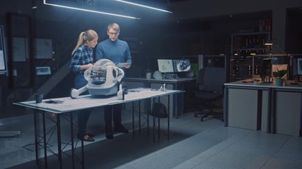 Poster - Late at Night in Robotics Engineering Facility Two Technical Engineers Talk and Work on a Wheeled Robot Prototype. In Background High Tech Research Center with Screens Showing Technology and Design