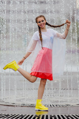 Young wet pretty girl with two braids in yellow boots and with transparent umbrella stands inside of fountain.