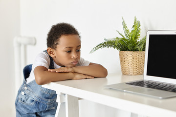 Wall Mural - People, childhood, technology and modern electronic gadgets. Upset sad African American schoolboy feeling bored, pouting lips, forbidden to use internet, looking at laptop with disappointment