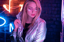 Beautiful Young Woman Wearing Metallic Bomber Jacket Trendy Stylish Glasses Stand Near Brick Wall Illuminated With Neon Lights, Pretty Fashion Girl In Modern Cloth Eyewear In Night Glow On Nightclub