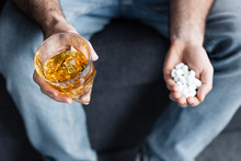 partial view of man holding glass of whiskey and handful of pills