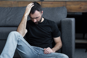 Wall Mural - depressed bearded man sitting on floor near grey sofa and holding hand on head