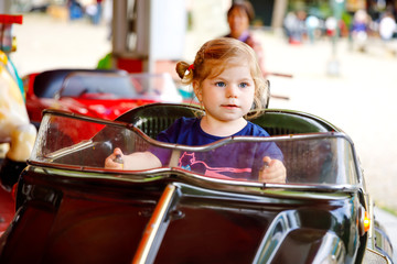 Wall Mural - Adorable little toddler girl riding on funny car on roundabout carousel in amusement park. Happy healthy baby child having fun outdoors on sunny day. Family weekend or vacations