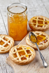 Fresh baked tarts with marmalade filling and apricot jam in glass jar on cutting board on rustic wooden table.