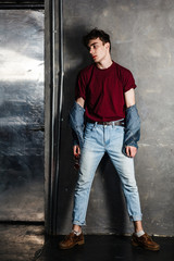 Full length portrait of stylish young fashion model man in bright red sunglasses and denim casual style posing near metallic door. standing and lean on grey wall and looking away. indoor, studio shot.