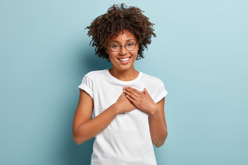 Indoor shot of happy dark skinned lady swears or promises something, holds hands on chest, tells truth, being honest, looks at camera friendly, has charming smile, wears casual white t shirt, glasses