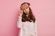 © wayhome.studio  - Displeased European woman wears bear sleep mask, pyjamas, tilts head, purses lips from negative feelings, has sleepy expression after awakening early in morning, isolated on pink background.