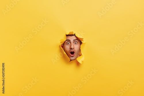 Fotografía Surprised young man with bristle keeps mouth widely opened, stares through torn paper wall, shows only face, expresses wonder and disbelief, poses against yellow background