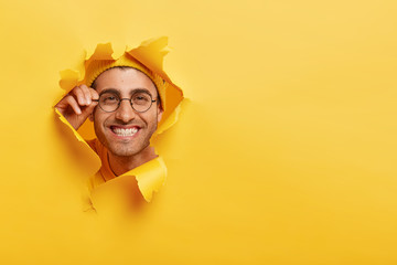 Close up shot of glad Caucasian man keeps hand on rim of spectacles, wears hat in one tone with background, looks happily through torn paper hole, copy space on right side for your promotional content