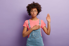 Confident Serious Woman With Curly Hair Makes Sincere Promise Or Oath, Keeps One Hand On Heart, Solemnly Swears, Raises Palm, Demonstrates Loyalty Gesture Being Honest Poses Against Purple Background.