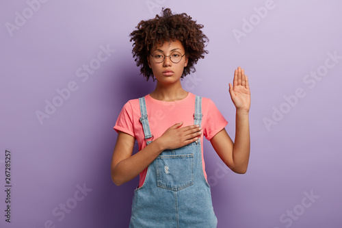 Confident serious woman with curly hair makes sincere promise or oath, keeps one hand on heart, solemnly swears, raises palm, demonstrates loyalty gesture being honest poses against purple background Tableau sur Toile