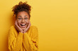 Sincere beautiful female laughs over funny joke, grins at camera with carefree expression, giggles as watches comedy, wears round spectacles and yellow sweater, stands indoor. Copy space for your text