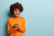 Photo of cheerful delighted African American woman types sms on modern cell phone device, enjoys good internet connection, dressed in orange jumper, focused aside, isolated on blue studio wall