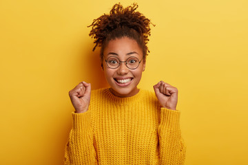 Happy woman with crisp hair, raises clenched fists, feels upbeat as gains goal, smiles broadly, has Afro hair, being fan of favourite team, wears optical glasses and yellow sweater, models indoor