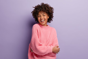 Wall Mural - Sincere gentle feminine model expresses positive emotions and feelings, being glad to pose on camera for making photo, dressed in oversized pink sweater, shows white teeth, has healthy skin.