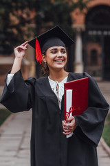 Wall Mural - Dreaming of a bright future. Thoughtful young woman in graduation gowns holding diploma and looking away