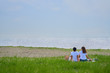 © Tak - 海岸で芝生の上に座る若いカップル、爽やかな夏の海。
