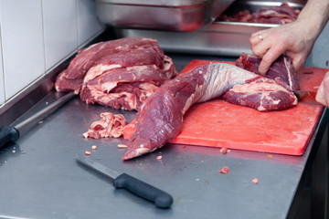 Closeup hands of man butcher chef cutting slices of fillet raw beef and pork meat off loin with knife in restaurant on red plastic board. Concept carve meat for barbeque grill on farmer market