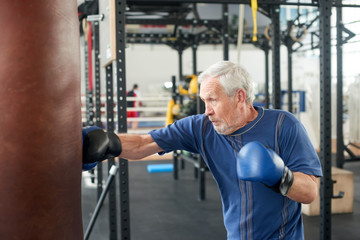 Elderly male person practicing box in gym. Healthy senior man in sportswear and blue gloves boxing punching bag in gym.