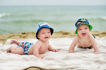 Half-year twins rest on the seafront. They lie on the bedspread on the sandy beach. Behind the back, see the surfboard. They are dressed in charming hats and bright shorts.