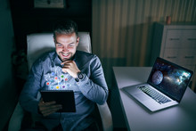 Young Smiling Caucasian Bearded Employee Using Tablet For Social Networks While Sitting In The Chair Late At Night. Next To Him Is Laptop.