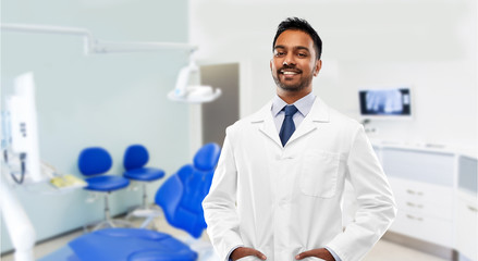 Wall Mural - medicine, dentistry and profession concept - smiling indian male dentist in white coat over dental clinic office background