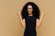Studio shot of overjoyed cheering woman with curly hair, raises clenched fists, celebrates success and triumph, dressed in casual black t shirt, eyewear, stands against brown wall with empty space