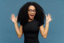 Slim Overemotive Dark Skinned Woman Raises Hands, Opens Mouth, Gestures Actively From Positive Emotions, Focused Down, Dressed In Casual Black T Shirt, Isolated Over Blue Background. Ethnicity Concept