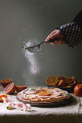 Young man's hand sprinkling sugar powder homemade Cheesecake with sicilian blood oranges, decorated by edible flowers, mint leaves served on grey linen table cloth with wall at background.