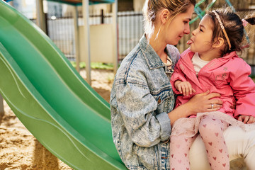 Candid outdoors image of funny cute little girl showing tongue to her mother, spending time together. Woman with her child playing at playground, feeling happy and loving each other.