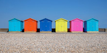 A Line Of Six Multicoloured Beach Huts On Seaford Beach, In The Foreground Is A Pebble Beach In The Back Ground Is A Clear Blue Sky