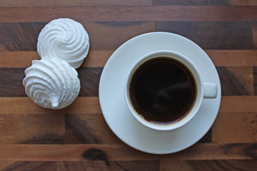 Black coffee in a white Cup on a saucer with two round white meringues on a brown wooden table