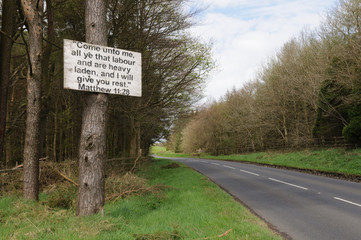 Wall Mural - Sign with a bible verse nailed to a tree by the side of an empty road, typical of the Northern Ireland countryside.