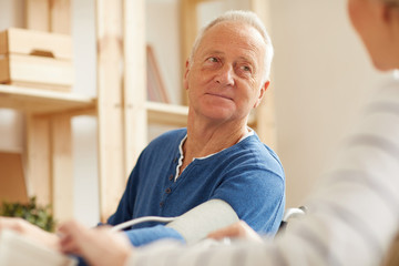 Low angle portrait of senior man checking his blood pressure with electric monitor at home lit by sunlight, copy space