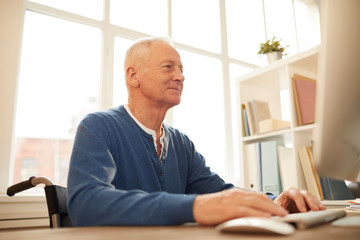 Low angle portrait of smiling senior man sitting in wheelchair using computer at home, copy space