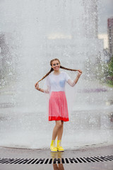 Canvas Print - Young wet pretty girl with two braids in yellow boots and with transparent umbrella stands inside of fountain.