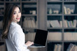 © NAMPIX - Portrait of young asian woman holding laptop and looking at camera while standing in library.