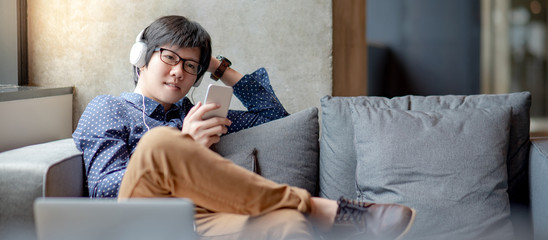 Wall Mural - Young Asian man using smartphone for listening to music on mobile application, sitting and relaxing on sofa during free time. Urban lifestyle in living space concept