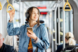 Fototapeta  - Young woman passenger standing with headphones and smartphone while moving in the modern tram, enjoying trip at the public transport