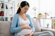 © PR Image Factory - worried pregnant woman sitting on sofa at home frowning face considering problem. asian girl with baby in belly sitting on couch upset sadness resting in living room indoors. stressed motherhood.