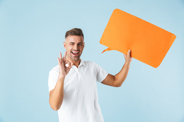 Poster - Excited emotional adult man posing isolated over blue wall background holding speech bubble.