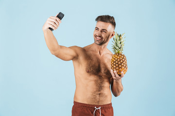 Poster - Excited happy adult man posing isolated over blue wall background take a selfie by phone holding pineapple.